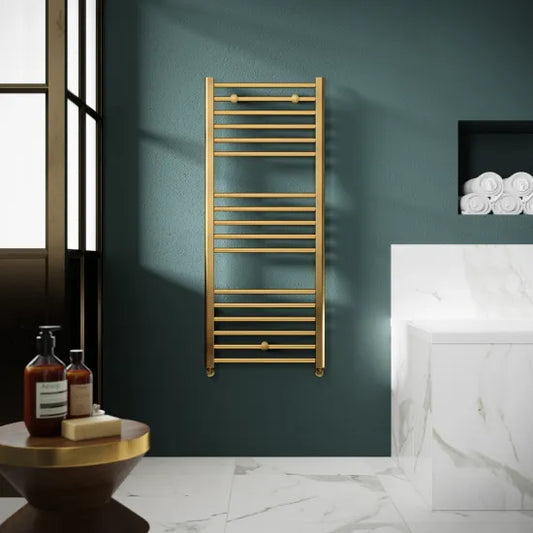 A brushed brass towel radiator mounted on a wall in a room with a dark green wall, a white marble floor, a round wooden side table, and a glass door.